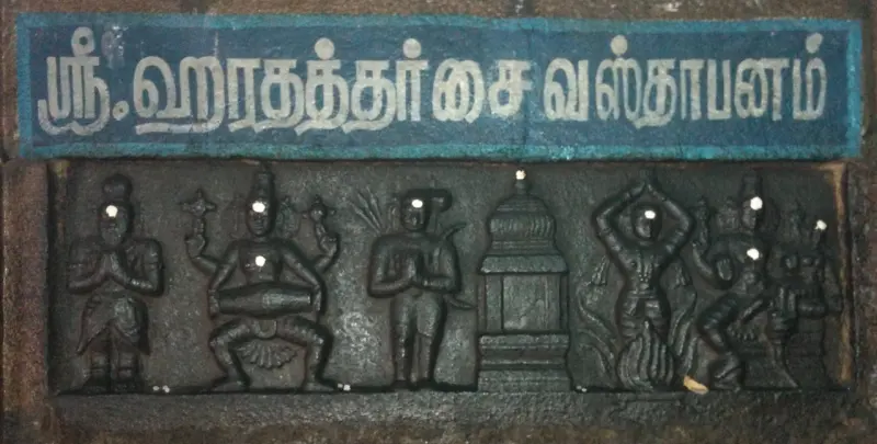 A dark stone relief of several carved figures beneath a blue board with Tamil lettering.
