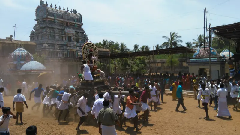 Many people haul a chariot with ropes across a sandy ground, with the temple tower and domes behind.