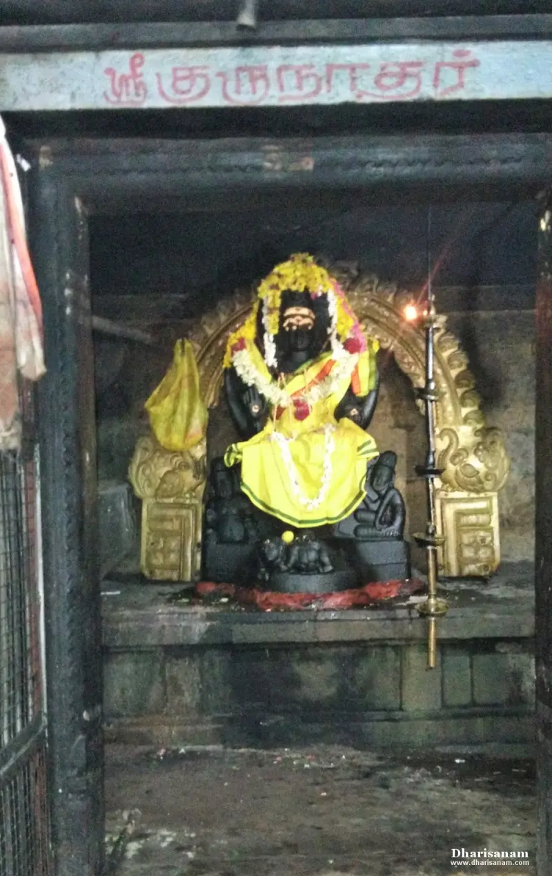 A dark stone Guru figure garlanded in yellow, seated in a shrine under a Tamil name board