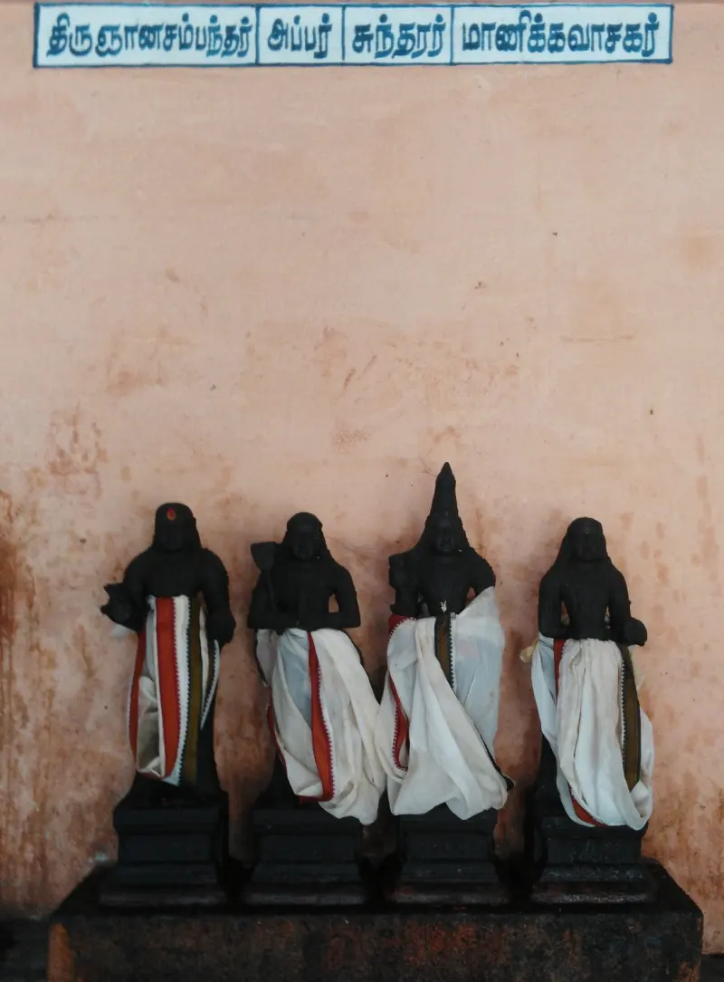 Four small dark idols in white cloth stand on a stone base beneath a board with their four names in Tamil.