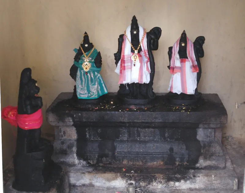 Three dark stone idols in cloth and a gold necklace stand on a platform, with a smaller kneeling figure at left.