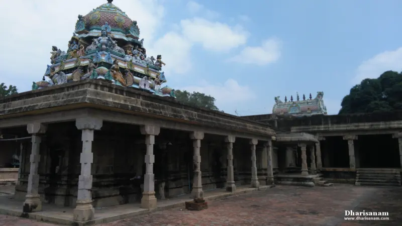 A stone pillared corridor with a painted tower rising above it, another shrine roof at right, under a blue sky