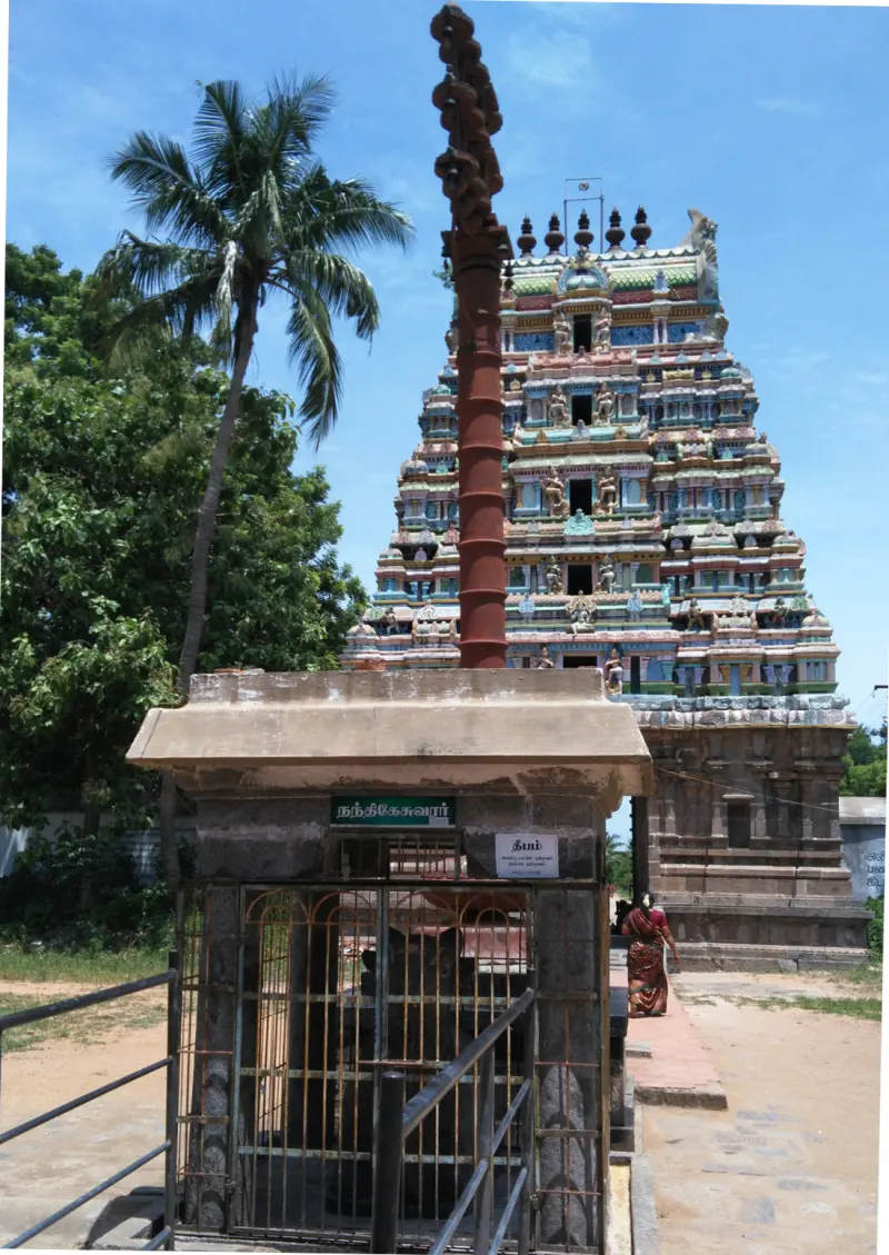 A tall flagstaff beside a small barred stone shrine with a Tamil sign; a colourful tower stands behind.