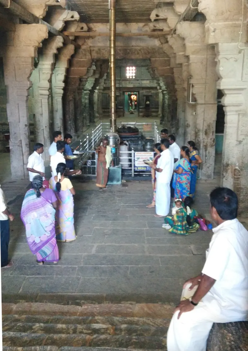 Devotees gather in a long pillared hall around a tall brass flag post, with a lit shrine far ahead.