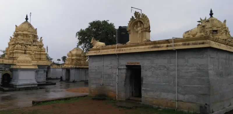 Golden-coloured shrine towers behind a grey stone shrine with a Nandi statue on its roof, under a cloudy sky.