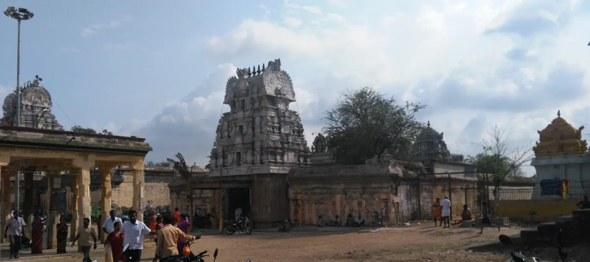 A stone tower and compound wall with a crowd in the foreground, a second shrine tower at the right and trees behind