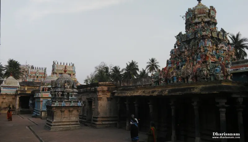 A sculpted tower rising behind a pillared hall, with small shrines and a few visitors on the paved ground