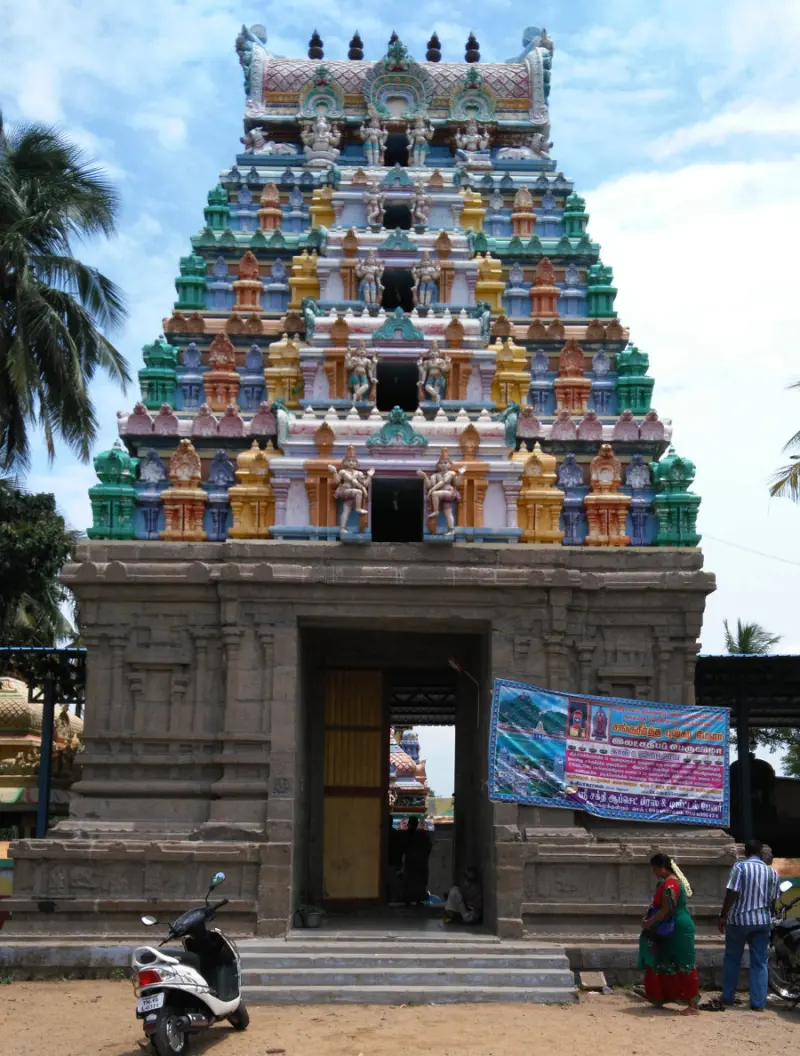A tall painted gopuram of five tiers rising over a stone gateway, with people standing before it.