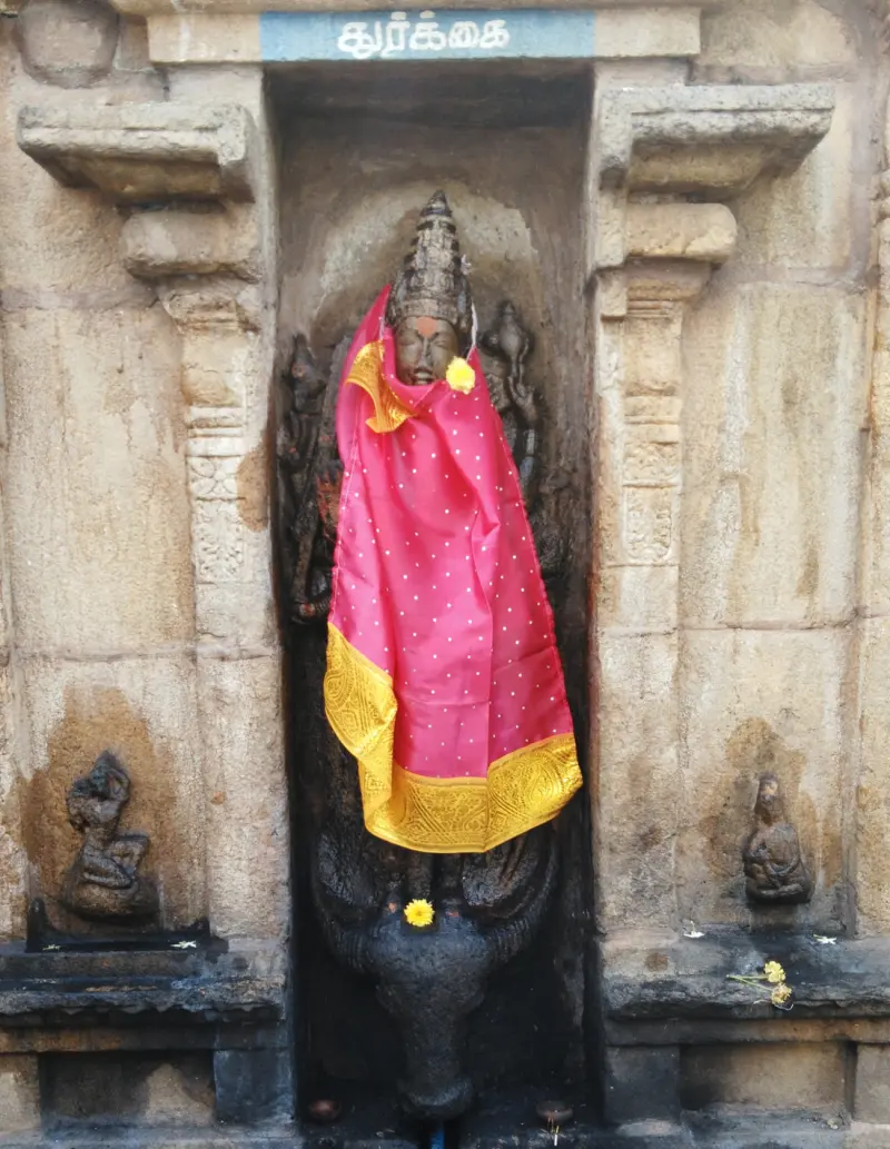 Stone idol in a wall niche, draped in a pink cloth with yellow flowers, under a Tamil name board