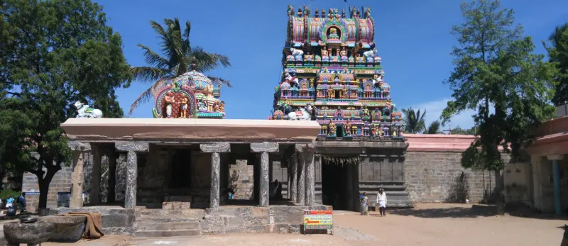 A stone pillared mandapam with a painted tower rising behind it and a domed shrine on the left.