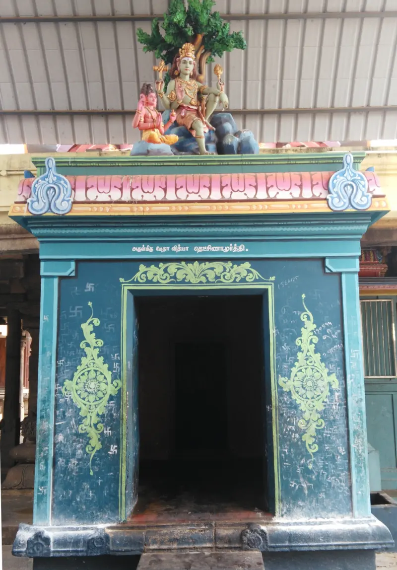 A dark blue shrine with floral patterns, a seated figure and a sage on top, and a Tamil sign naming Dakshinamurthy