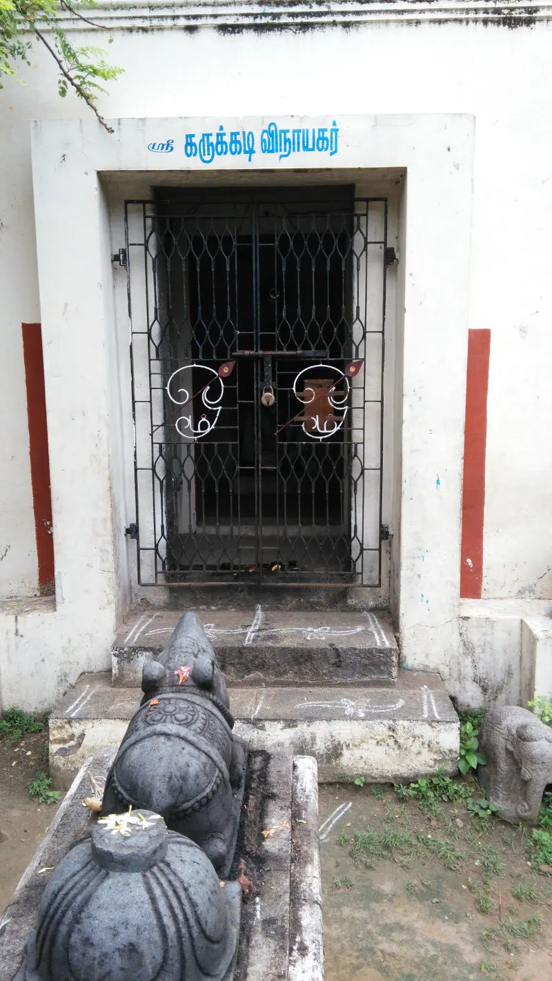 A white shrine with a barred iron gate and a name board in Tamil, a stone Nandi on a platform in front.