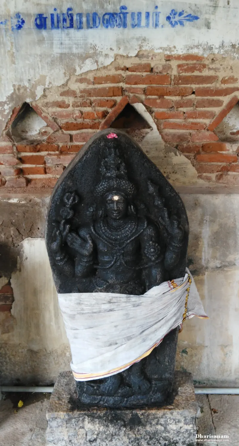 A black stone idol with several arms in a white cloth before a brick wall; a Tamil label above names Subramaniyar.