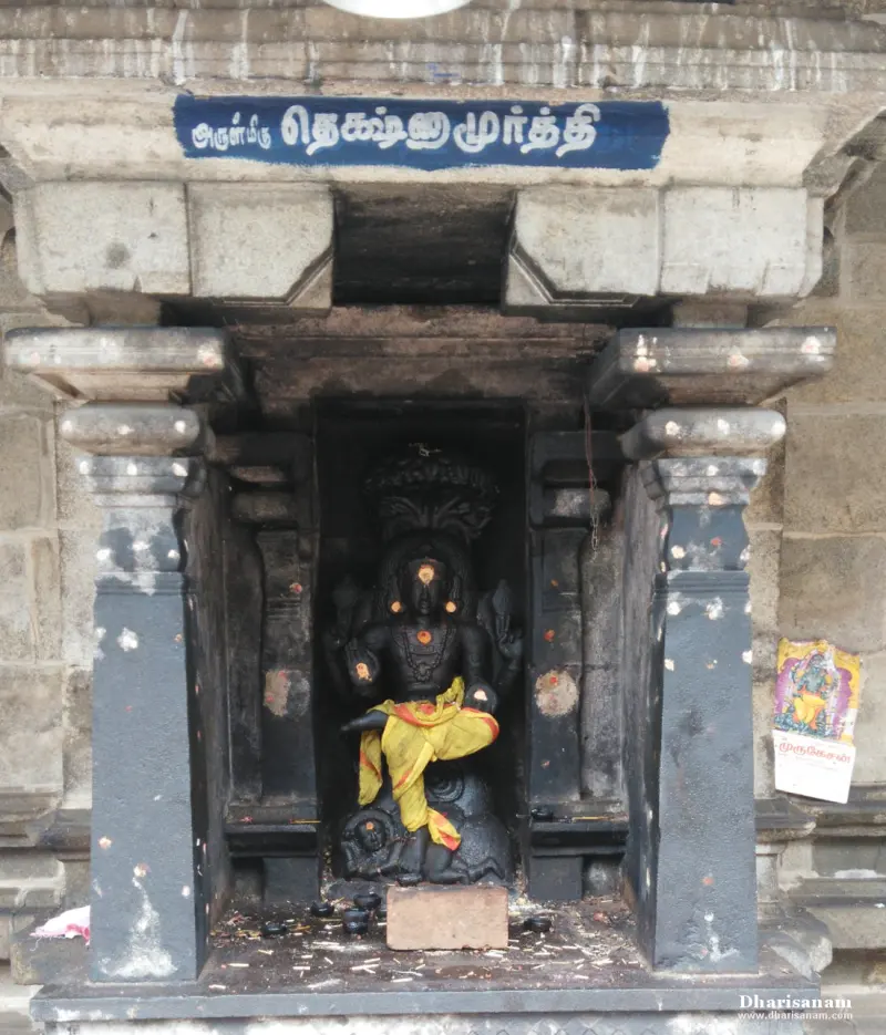 A dark stone Dakshinamurthy seated in a niche with a yellow cloth and a blue board naming him above