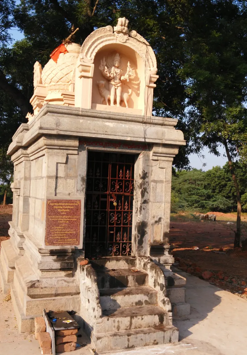 A small stone shrine with steps and an iron gate, a sculpted deity in an arch above, and trees behind.