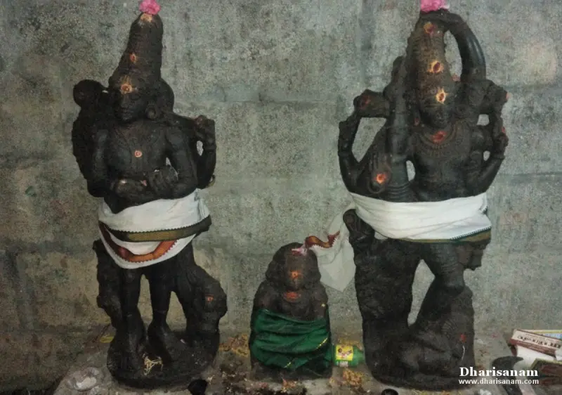 Two tall black idols in white cloth with a small idol between them, against a stone wall.