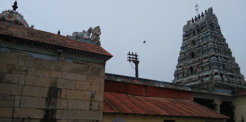 Tiled temple roof and a tall carved tower rising behind a stone wall under a grey sky.