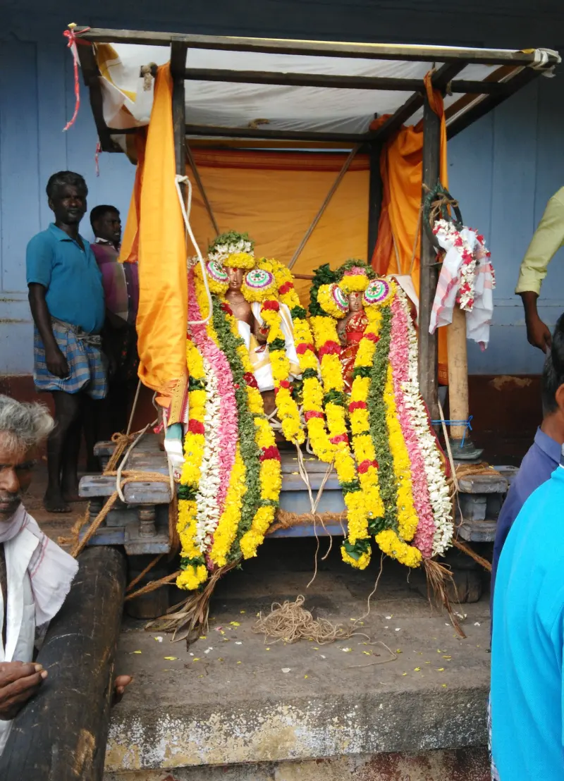 Two idols covered in flower garlands on a wooden carrier, with devotees around