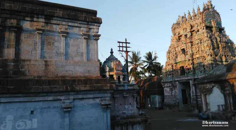A tall carved gopuram on the right, a flagstaff and a small domed shrine in the middle, and stone walls at left