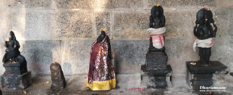 Four idols on stone bases, one in patterned silk, stand against a large-block stone wall beside a small serpent stone
