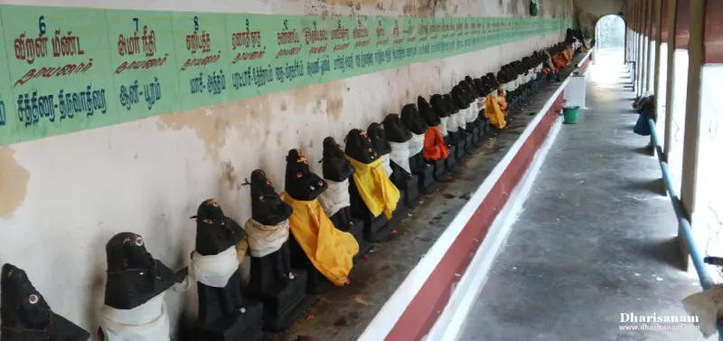 Many dark idols with cloth wraps stand in a line below a green board with Tamil names, beside an open corridor