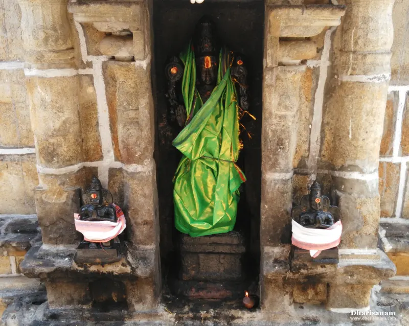 A dark stone figure draped in green silk stands in a niche between stone pillars, with two small figures at its feet
