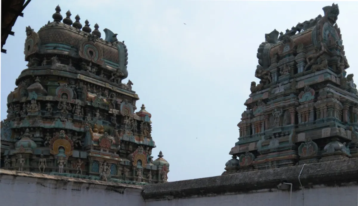 Two colourful towers covered in painted figures rise above a plain white wall against a pale sky.