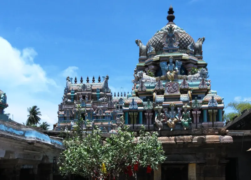Two colourful towers with sculpted figures against a blue sky; a leafy tree hung with red cloth in front.