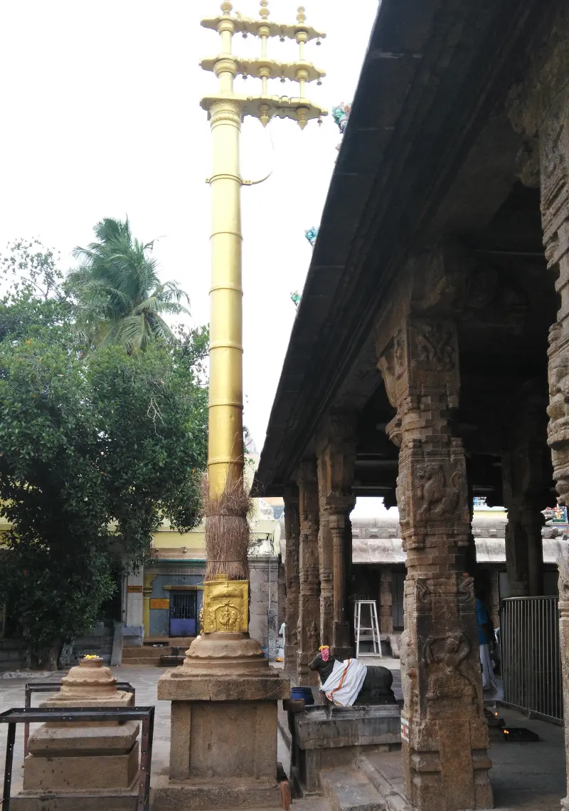 A tall golden flag post beside a hall of carved stone pillars, with a Nandhi nearby
