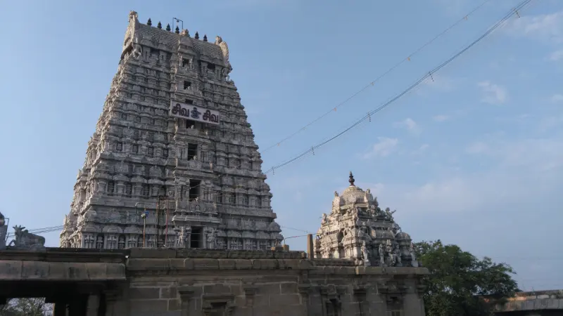 A tall tiered gopuram with a Tamil sign and a smaller carved tower, under a clear blue sky