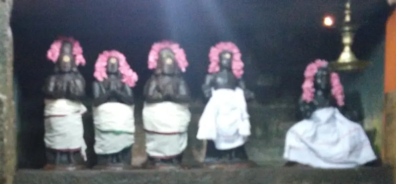Five small dark idols with pink flower garlands on their heads, dressed in white cloth, in a dim shrine