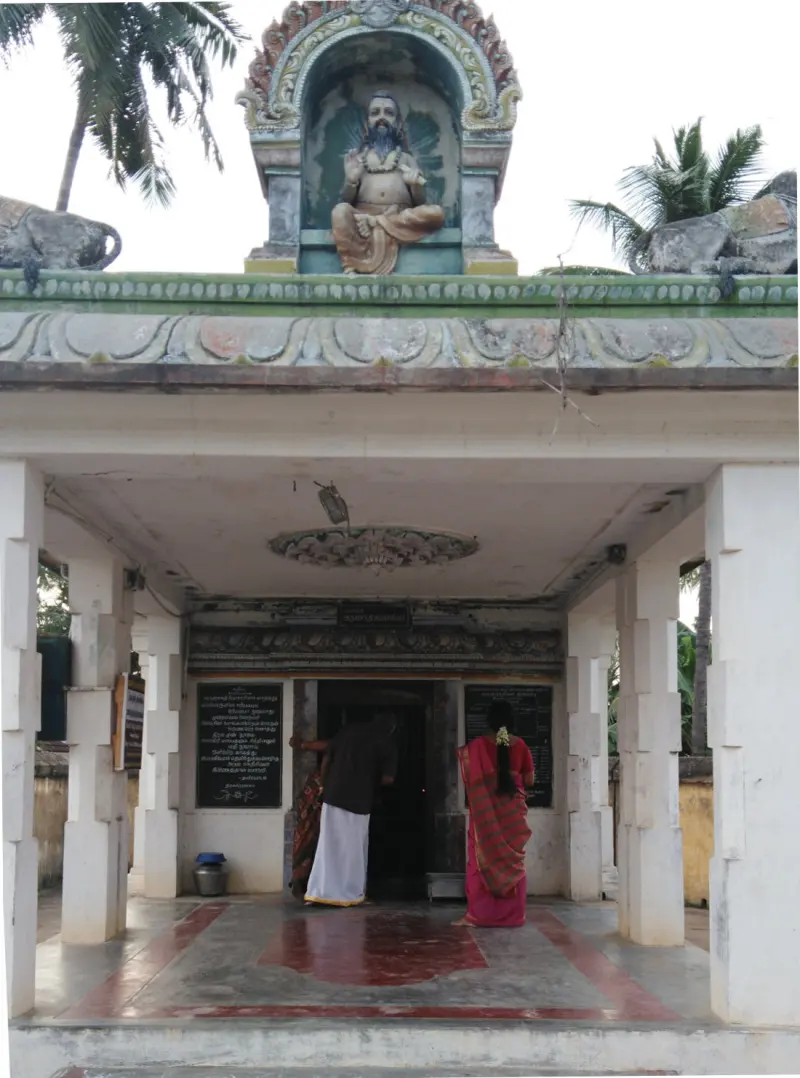 A white pillared porch with a seated bearded figure above it, and two devotees at the dark doorway