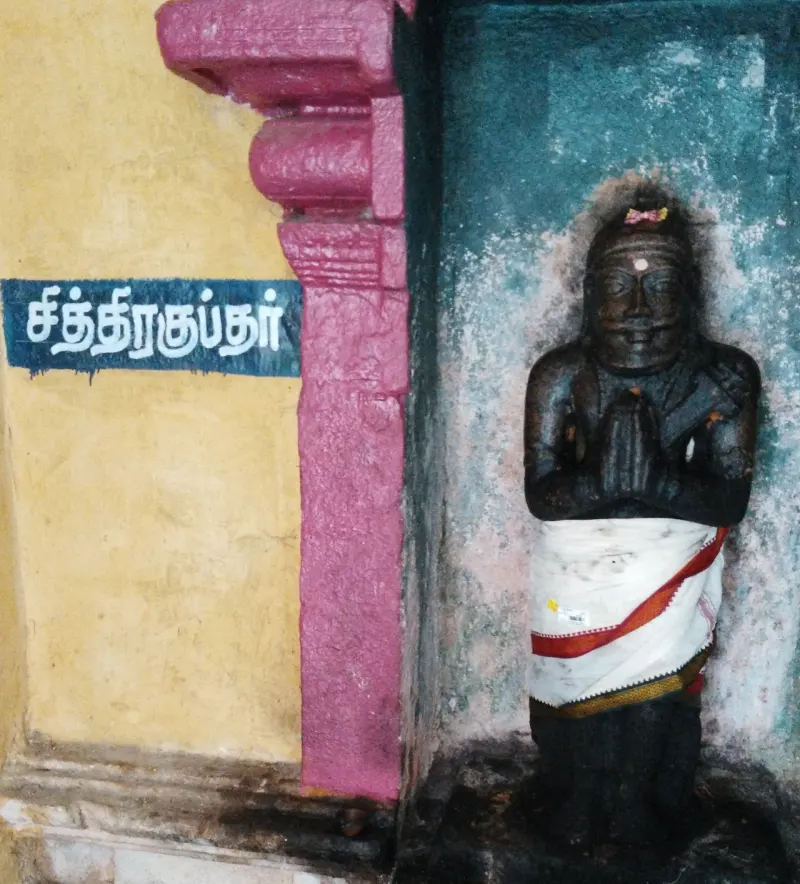 A dark stone idol with folded hands in white cloth, beside a blue board with the Tamil name Chitragupta.