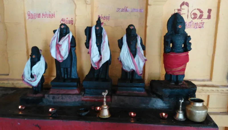 Five black stone idols in a row with lit lamps and a bell in front, and Tamil names painted on the wall behind