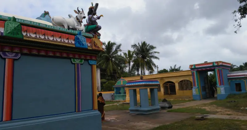 A blue shrine wall with a white bull on its roof; the entrance arch and a small mandapam beyond