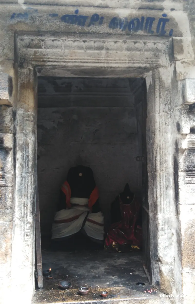 A narrow stone shrine with a Tamil board above, two dressed idols inside and small lamps in front