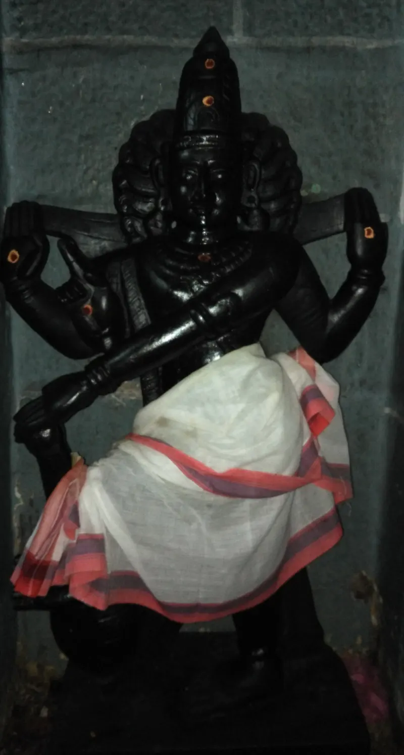 A four-armed black stone idol with one raised leg, wearing a white cloth with a red border, against a grey wall.