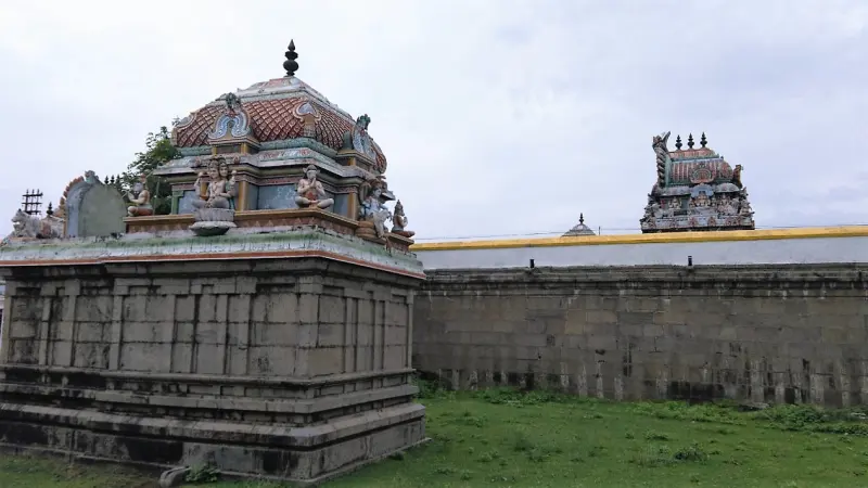 A small stone shrine with a domed roof and seated figures, a painted tower rising behind the wall.