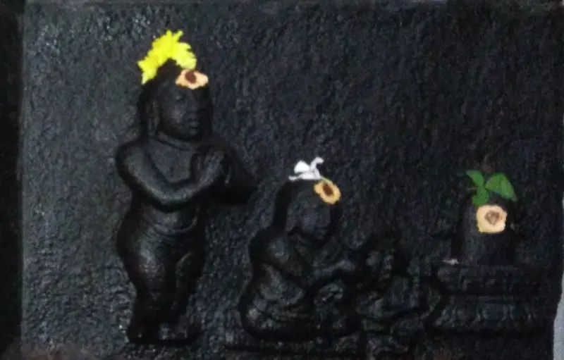A black stone relief with a standing figure with folded hands, a seated figure, and a lingam topped with green leaves